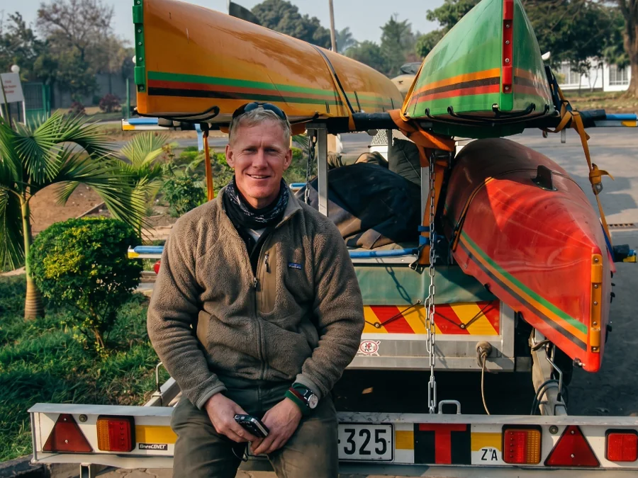 Tim Cook sitting on the RowZambezi boat trailer in Zambia, with the triple sculls loaded behind him
